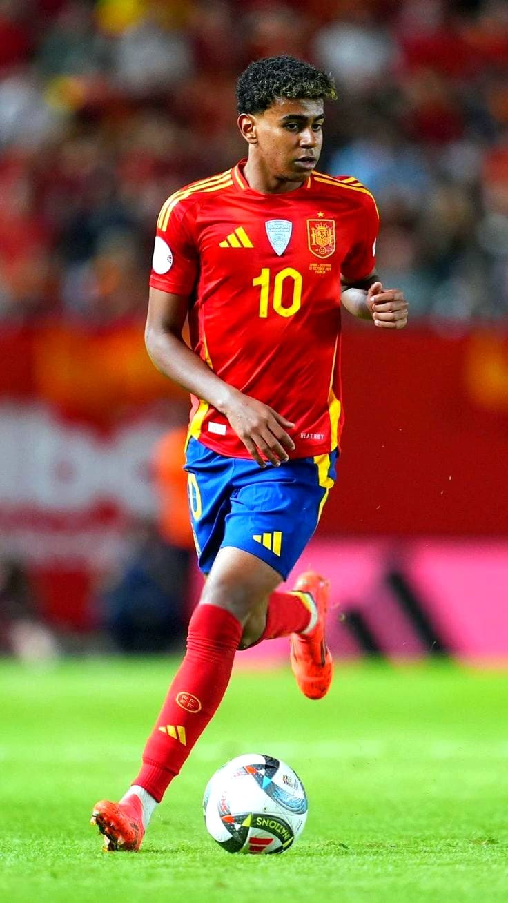 A male soccer player in a red and yellow Spain jersey with the number 10 dribbles a ball on the field during a match, broadcast live on IPTV Latino. The background shows a blurred crowd.