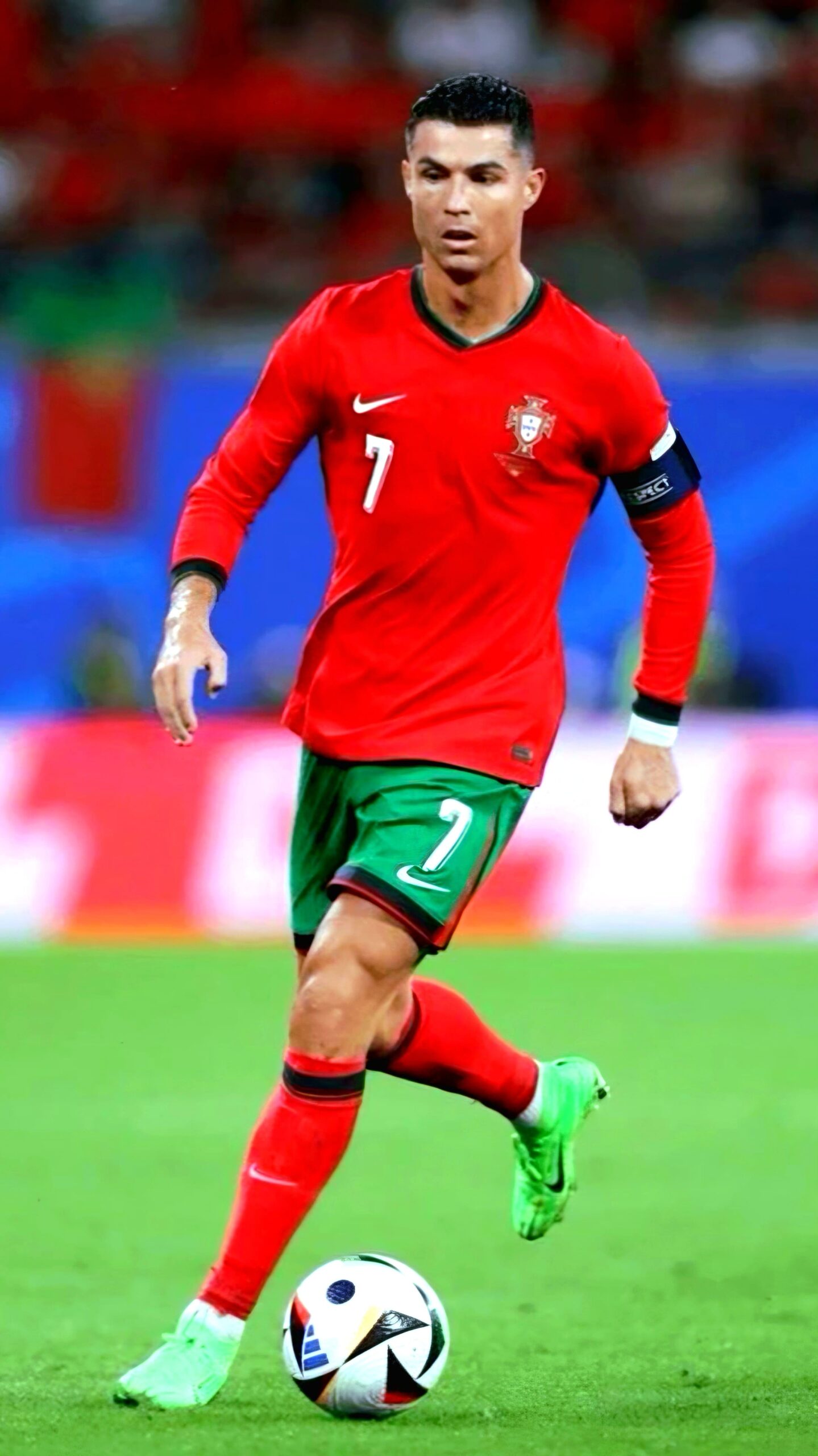 A soccer player in a red uniform with green shorts and neon green cleats is running on a field, focused on a soccer ball. Wearing number 7 and a captain's armband, he’s captured mid-play—perfect for streaming on IPTV Latino with the crowd blurred behind him.